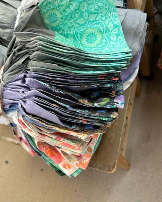 A large stack of folded patterned fabric pieces in various colors and designs, including green, purple, blue, and floral prints, piled on a wooden table.