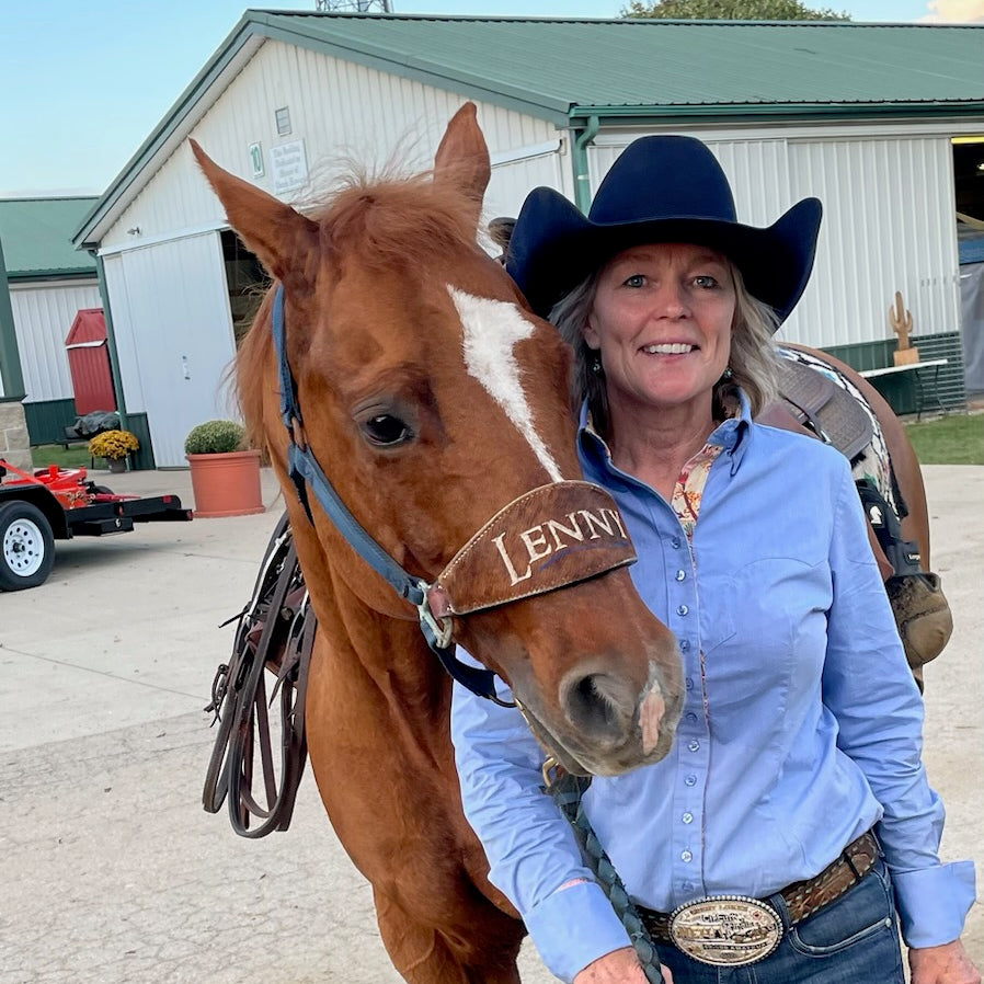 A smiling woman in a cowboy hat and blue shirt stands beside a brown horse wearing a branded halter that reads 'LENNIE'. The setting is outdoors in front of a white barn with a green roof.