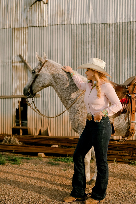 Woman in a pink western shirt, hat, jeans, belt, and boots standing beside a gray horse