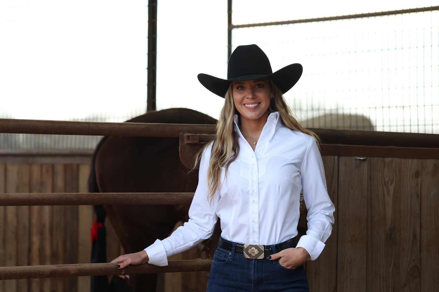 A smiling woman wearing a black cowboy hat, white button-down shirt, and dark blue jeans stands in a rustic barn setting with a wooden fence and a horse behind her.