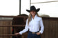 A smiling woman wearing a black cowboy hat, white button-down shirt, and dark blue jeans stands in a rustic barn setting with a wooden fence and a horse behind her.