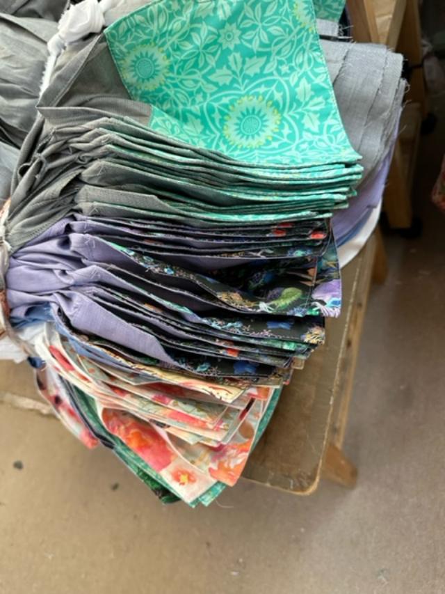 A large stack of folded patterned fabric pieces in various colors and designs, including green, purple, blue, and floral prints, piled on a wooden table.