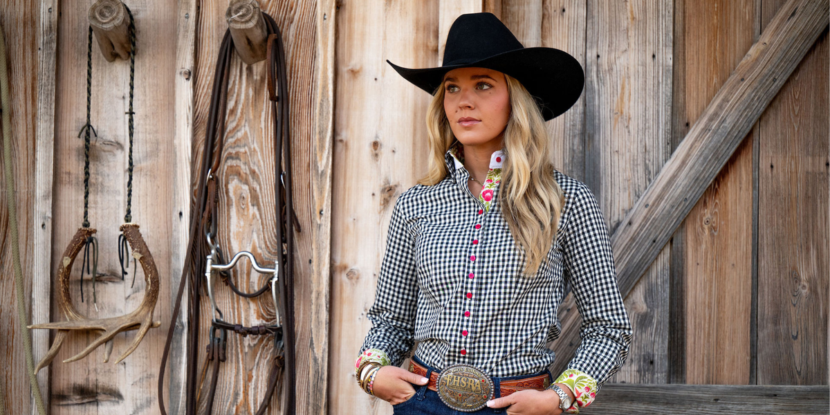 A woman wearing a black cowboy hat, a black-and-white gingham western shirt with colorful embroidery, and a large silver belt buckle stands in front of a rustic wooden wall with hanging leather tack.