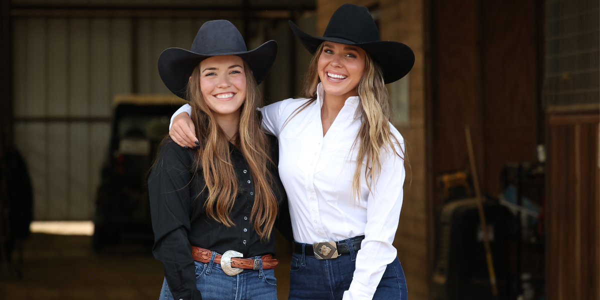 Two women standing together in front of a rustic barn, wearing cowboy hats and western-style shirts. One wears a black long-sleeve button-up shirt with a brown belt, and the other wears a white long-sleeve button-up shirt with a dark belt.