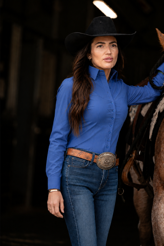 A woman wearing a bright blue long-sleeve western-style button-up shirt, blue jeans, a brown leather belt with a large silver buckle, and a black cowboy hat, standing next to a horse in a dimly lit indoor setting.