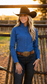 A woman wearing a bright blue long-sleeve western-style button-up shirt, dark jeans, a brown leather belt with a large decorative buckle, and a black cowboy hat, standing in front of a rustic wooden fence during golden hour.