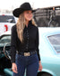 A woman wearing a black cowboy hat, black long-sleeve button-up shirt, and dark blue jeans stands outdoors next to a light blue vintage car. She is wearing a black belt with a large decorative buckle.