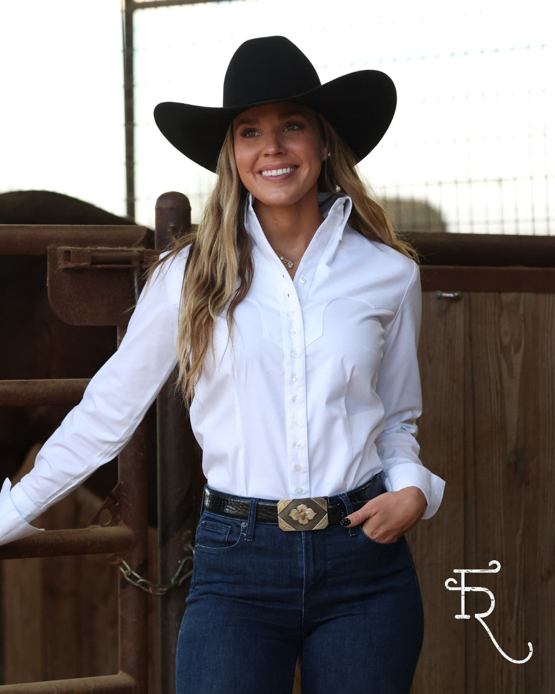 A woman wearing a black cowboy hat, a white long-sleeve button-down shirt, and dark blue jeans with a western belt stands in front of a wooden fence.