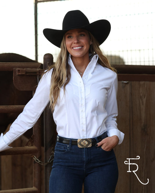 A woman wearing a black cowboy hat, a white long-sleeve button-down shirt, and dark blue jeans with a western belt stands in front of a wooden fence.
