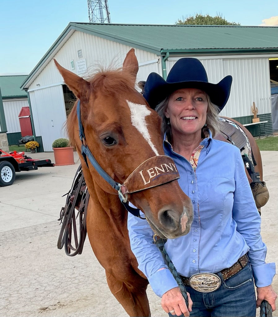 A smiling woman in a cowboy hat and blue shirt stands beside a brown horse wearing a branded halter that reads 'LENNIE'. The setting is outdoors in front of a white barn with a green roof.